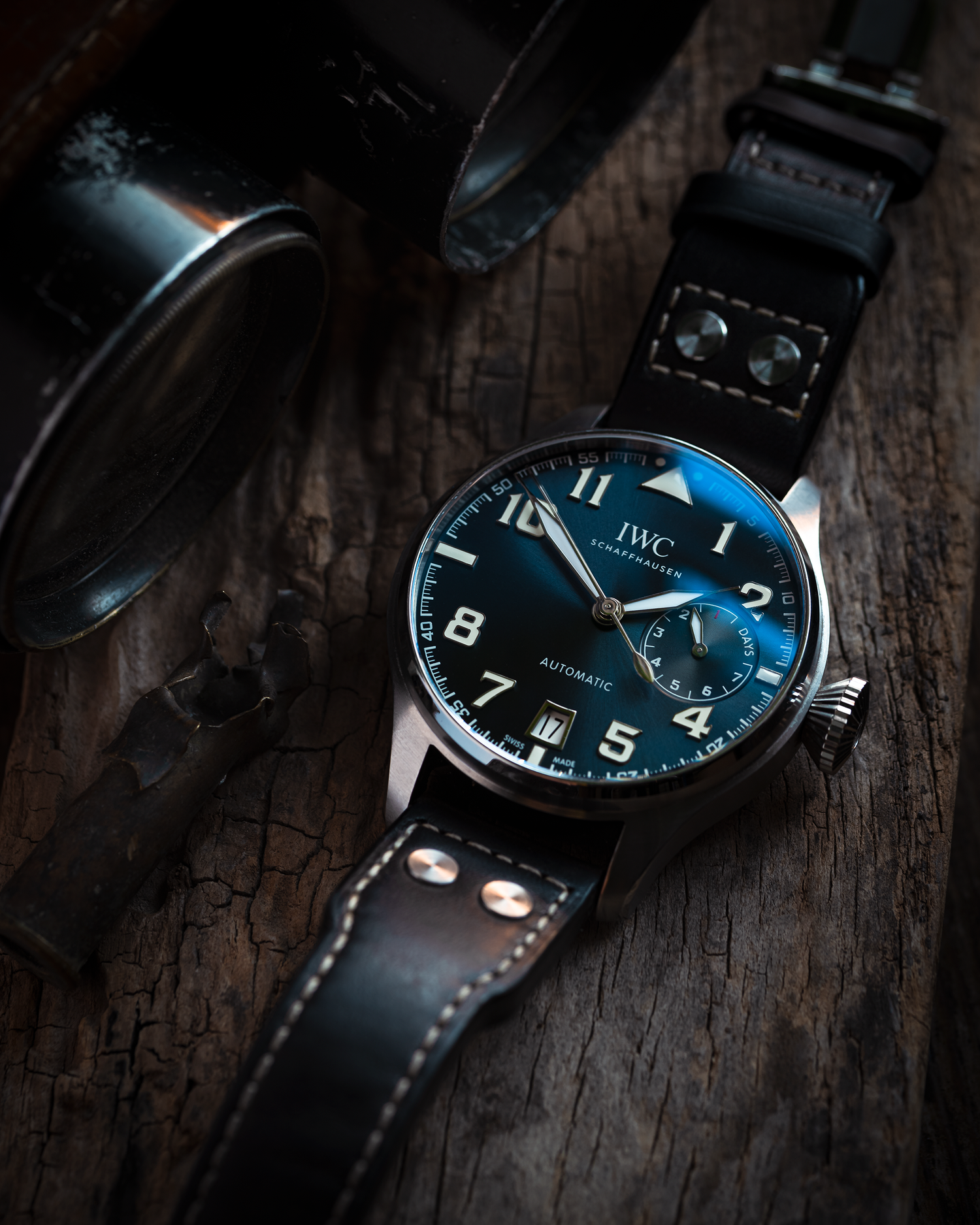 A close-up photograph of an IWC Big Pilot watch placed on a wooden board. The watch is accompanied by binoculars and an empty gun cartridge, serving as props.