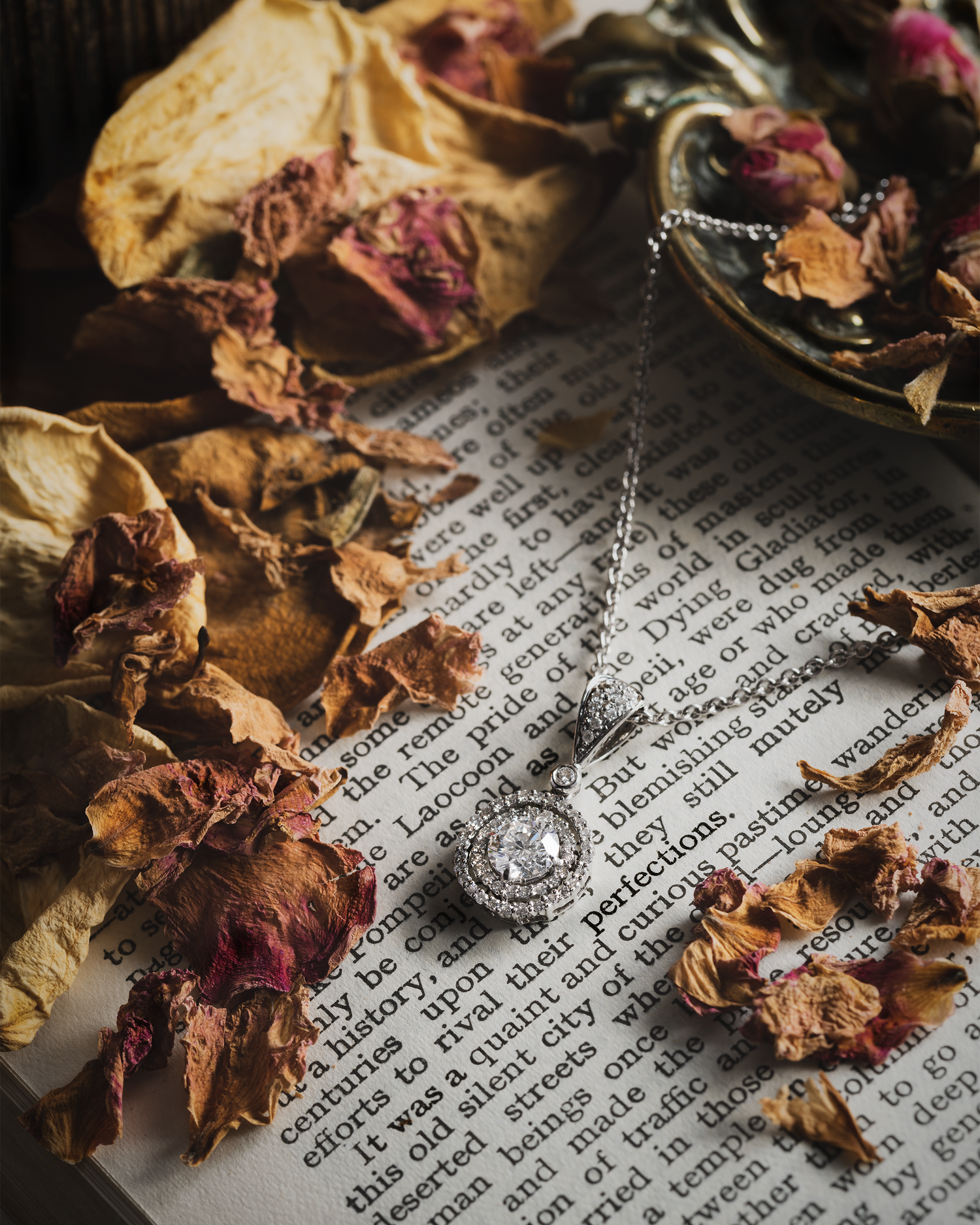 A diamond halo necklace photographed on the pages of a book with dried petals 