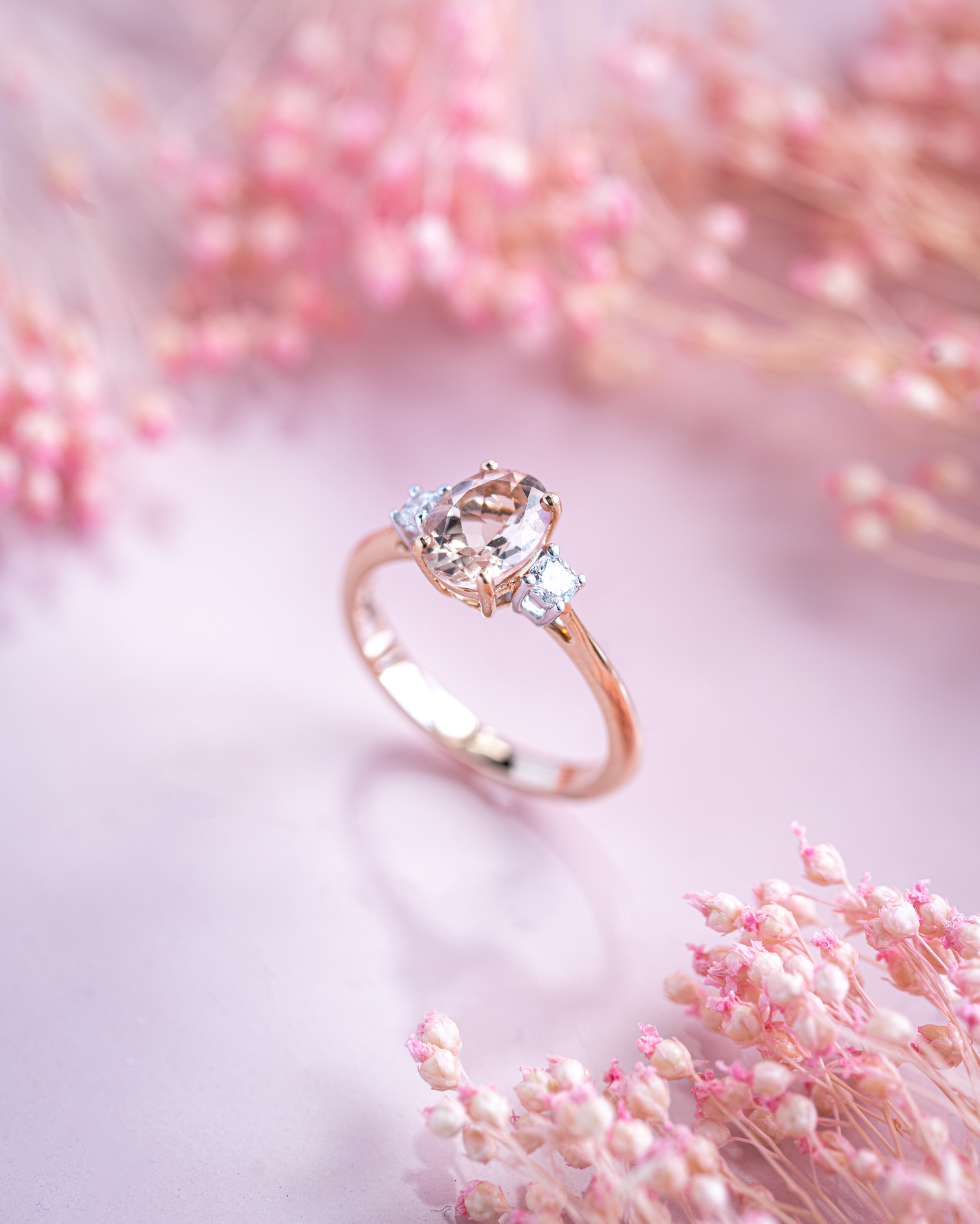 A oval morganite set in a simple four claw setting with two diamonds either side. Photographed on a pink base with pink flowers to accompany.