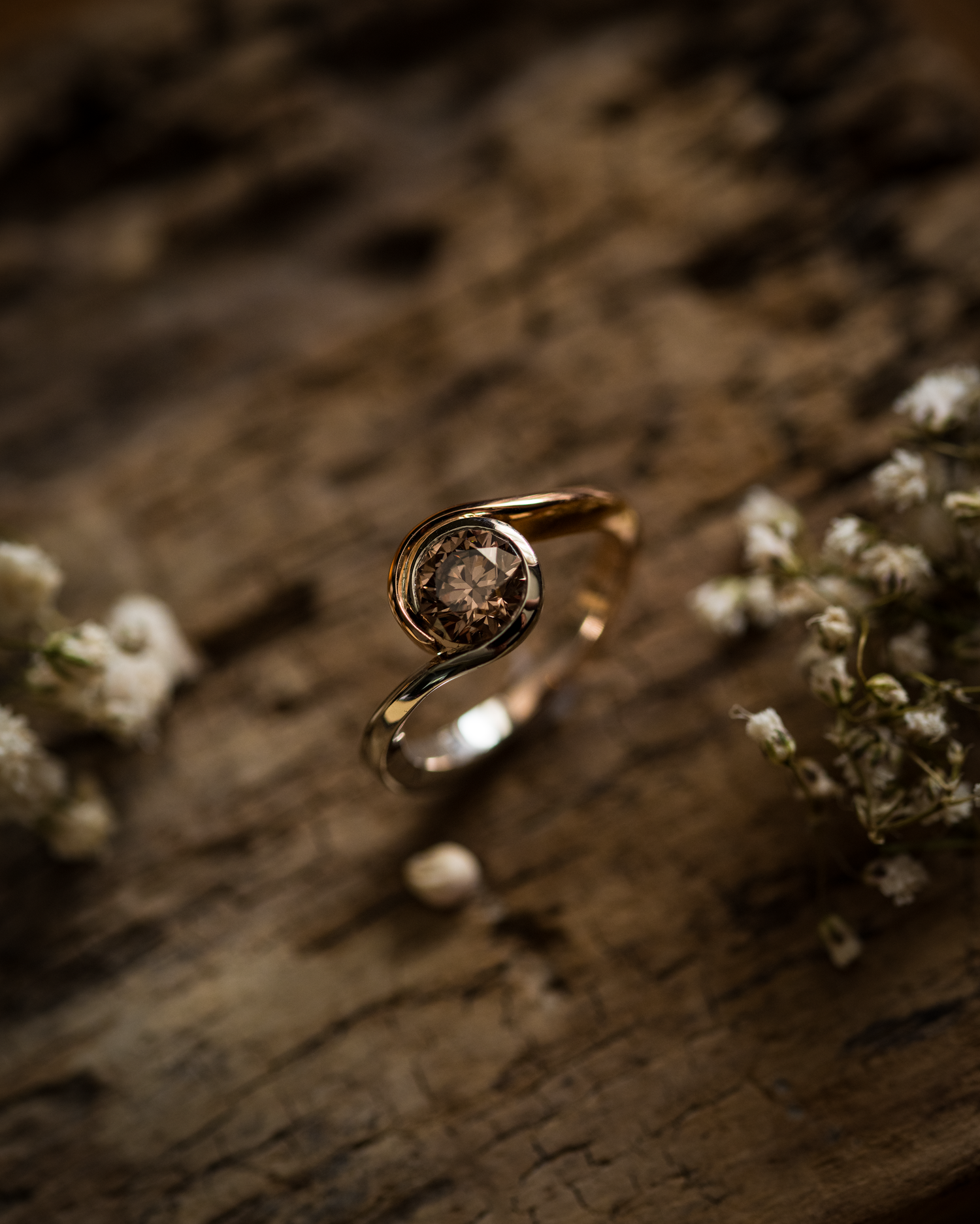 The pink and brown diamond ring sits within a twist design and is photographed on a wooden background with dried flowers to accompany the composition