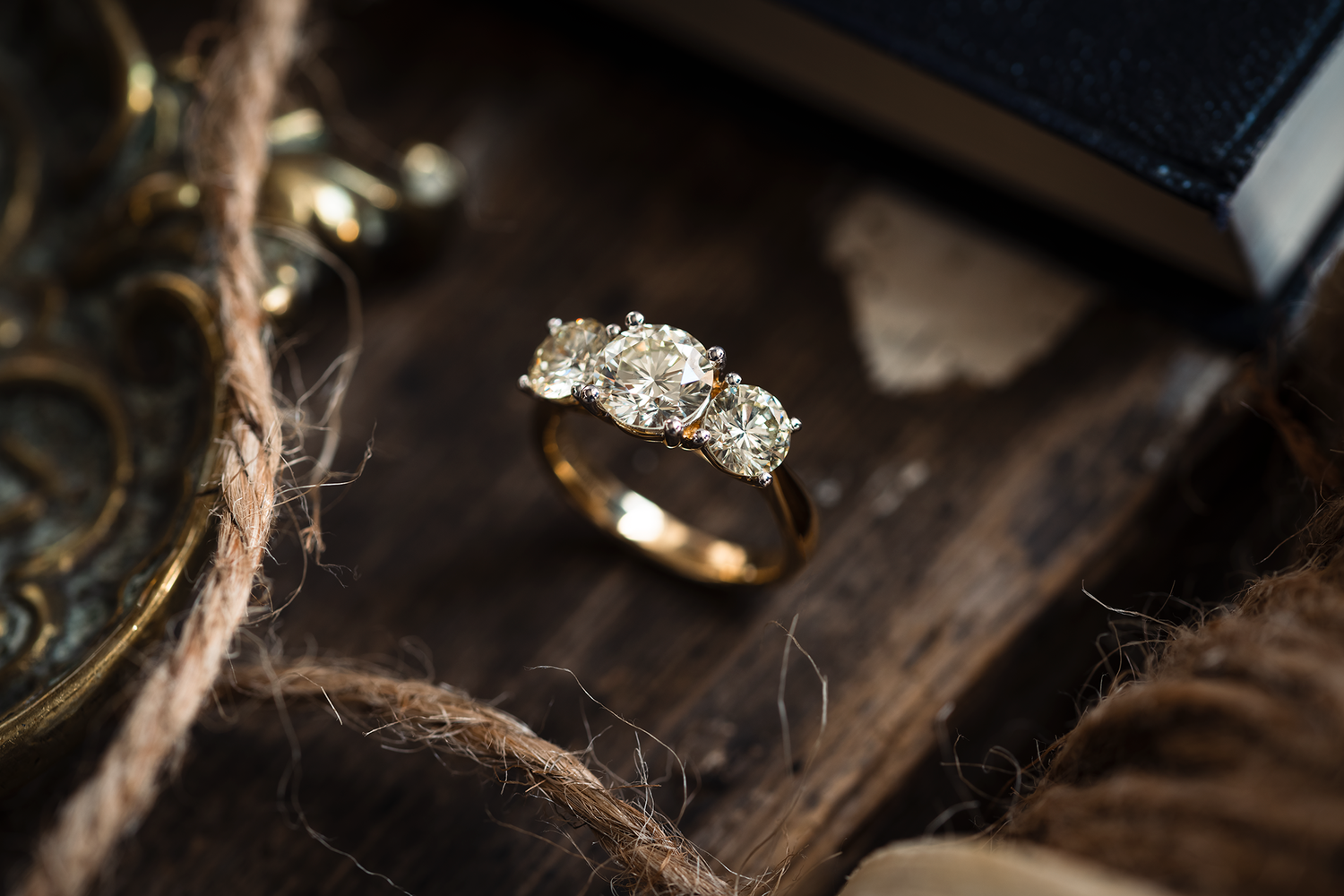 A three stone vintage diamond ring photographed on a wooden board complete with string, the corner of a book and hints of antiques to complement the image.
